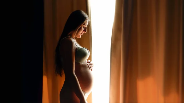 Krásná těhotná žena v lingeri otevírá oponu na velké okno a dívá se na její velké břicho. — Stock fotografie