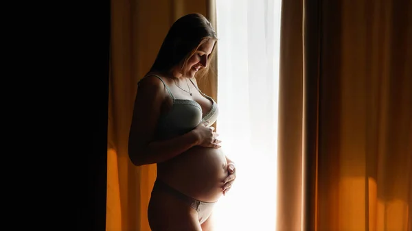 Krásná usměvavá těhotná žena rozhrnuje závěsy a stojí u velkého okna v hotelovém pokoji. Koncept šťastného těhotenství a očekávání dítěte — Stock fotografie