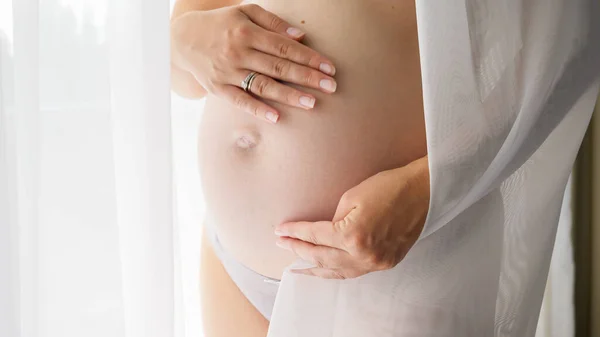 Close-up de mulher grávida cobrindo em tecido branco e segurando sua grande barriga crescente. Conceito de gravidez feliz e antecipação do bebê — Fotografia de Stock