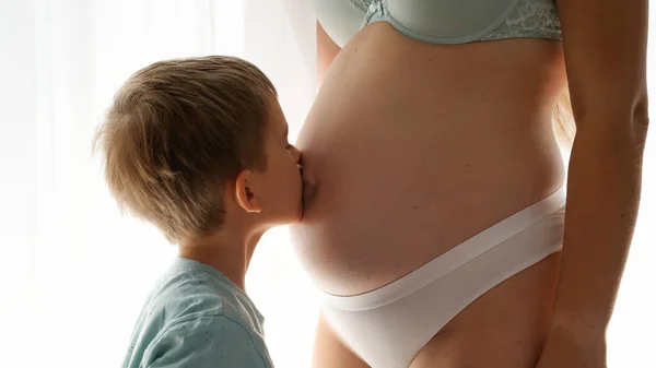 Крупный план маленького мальчика, целующего свою беременную маму в большом животе, стоящего у окна в спальне. Концепция семейного счастья и предвкушения ребенка — стоковое фото
