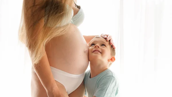 Joven madre embarazada abrazando y acariciando a su pequeño hijo abrazándola en la gran ventana del dormitorio. Concepto de felicidad familiar, amor y anticipación del bebé — Foto de Stock