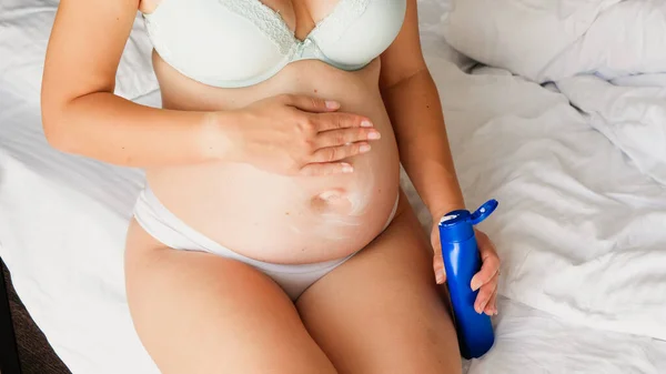 Toned image of pregnant woman sitting on bed and applying moisturizing creme or lotion — Stock Photo, Image