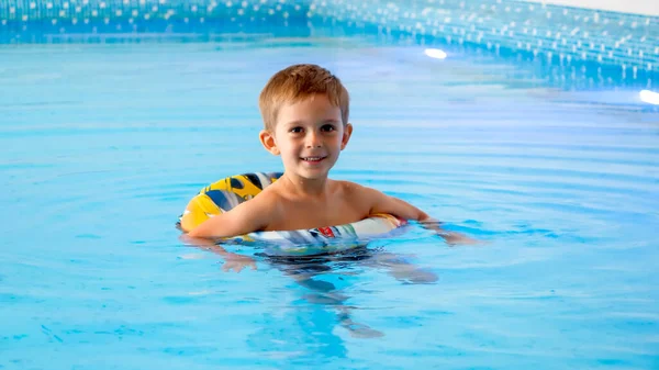 Feliz niño alegre usando el anillo inflable mientras que aprende a nadar en piscina cubierta en el hotel. —  Fotos de Stock