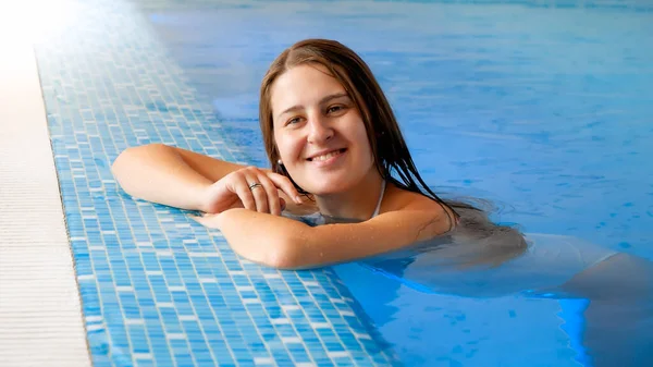 Retrato de una hermosa morena sonriente apoyada en el lado de la piscina en el spa del complejo hotelero y mirando en cámara — Foto de Stock