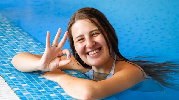 Mutlu gülümseyen esmer kadının portresi yüzme havuzunda dinleniyor ve parmaklarıyla işaret veriyor.. — Stok fotoğraf