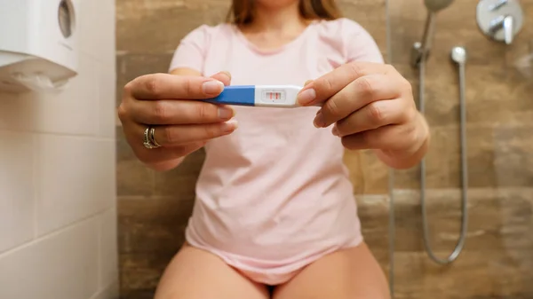 Крупный план молодой беременной женщины с положительным тестом на беременность с двумя полосками в камере, сидя на унитазе — стоковое фото