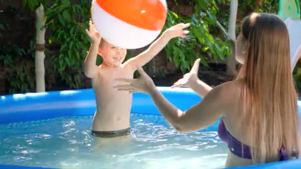 Glücklich lächelnder und lachender kleiner Junge, der seiner Mutter im Schwimmbad einen bunten Strandball zuwirft. Sommerurlaub und Urlaub für Familien — Stockvideo