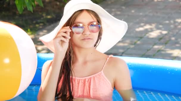 여름 모자를 쓰고 멋지게 웃는 십 대 소녀와 집 뒤 뜰에 있는 푹신 한 수영장에서 편안하게 쉬고 있는 멋진 선글라스. 행복 한 여름 휴가와 휴가의 개념 — 비디오