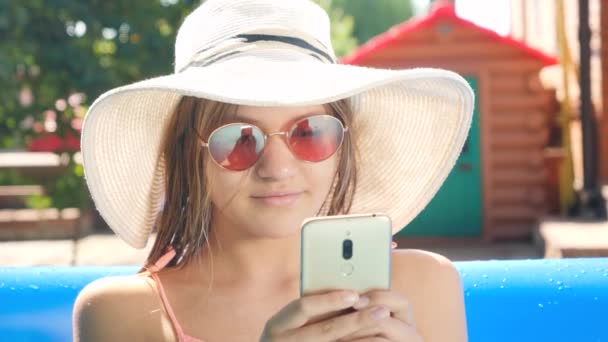 Retrato de menina sorridente em chapéu e óculos de sol relaxando na piscina e usando smartphone. Conceito de férias felizes de verão e férias — Vídeo de Stock