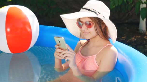 Ritratto di bella ragazza che si rilassa nella piscina all'aperto nel cortile e digita il messaggio sullo smartphone. Concetto di buone vacanze estive e vacanze — Video Stock