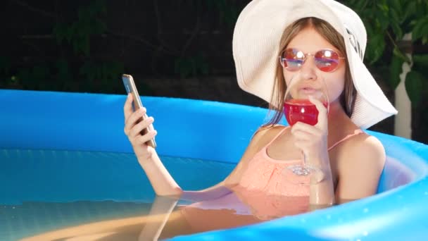 여름 모자를 쓴 소녀의 아름다운 미소와 선글라스는 스마트폰으로 소셜 미디어를 둘러보면서 수영장에서 휴식을 취하고 칵테일을 마시는 것이다. 행복 한 여름휴가와 휴가의 개념 — 비디오