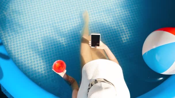 Horní pohled na mladou ženu relaxující a ležící v bazénu a pití koktejlu při použití smartphonu. Koncept šťastné letní dovolené a dovolené — Stock video