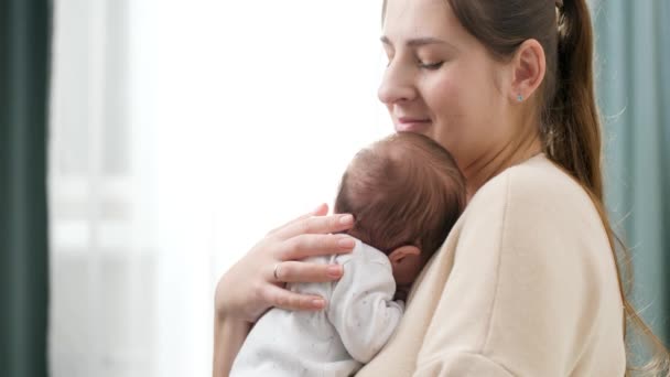 Gülümseyen genç bir kadının portresi. Yeni doğmuş bebeğini kucaklıyor ve yatak odasının penceresine yaslıyor. Aile mutluluğu ve küçük çocuklu sevgi dolu ebeveynler. — Stok video