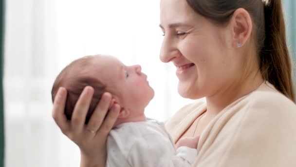 Retrato de primer plano de adorable bebé y madre sonriente mirándose unos a otros contra una gran ventana en la casa. Concepto de felicidad familiar y padres amorosos con niños pequeños — Vídeos de Stock