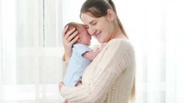 Güzel gülümseyen bir annenin portresi yeni doğmuş bebeğini parlak sabah güneşine karşı büyük bir pencerede okşuyor. Aile mutluluğu ve küçük çocuklu sevgi dolu ebeveynler.