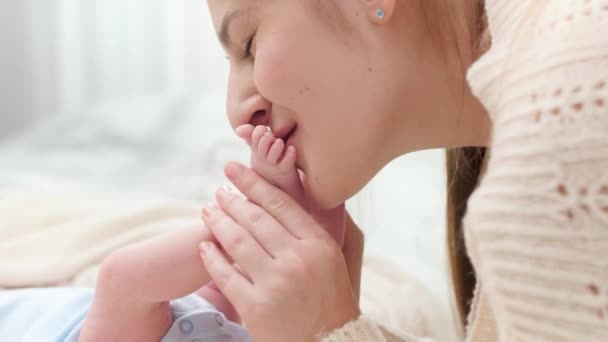 Primer plano de madre cariñosa y cariñosa sosteniendo y besando pequeños pies de su bebé recién nacido en la cama. Concepto de felicidad familiar y padres amorosos con niños pequeños — Vídeo de stock