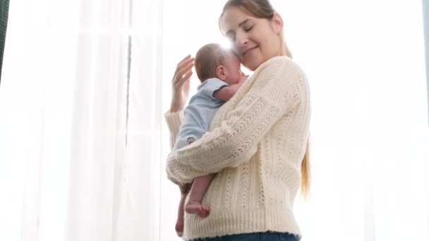 Szczęśliwa uśmiechnięta młoda matka przytula i całuje swojego nowonarodzonego synka przeciwko jasnemu słońcu świecącemu przez okno. Pojęcie szczęścia rodzinnego i kochających rodziców z małymi dziećmi — Wideo stockowe