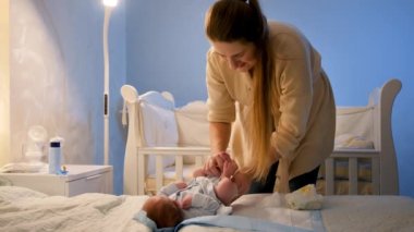 Gülümseyen genç bir anne bebek bezi değiştiriyor ve yeni doğan bebeğini siliyor. Geceleri yatakta çok dağınık oluyordu. Ailelerin ve bebek hijyeninin uykusuz geceleri