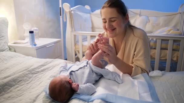 Feliz joven madre sonriente mirando a su bebé recién nacido acostado en la cama por la noche y besando sus pies. Concepto de felicidad familiar y paternidad. — Vídeo de stock