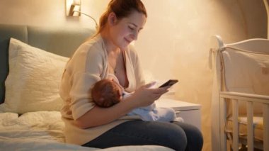 Gülümseyen genç anne, bebeğini anne sütüyle besliyor ve gece yatak odasında akıllı telefondan internette geziniyor. Sağlıklı ve doğal bebek beslenmesi kavramı. Annenin sağlığı ve...