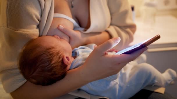 Fechar a mamãe alimentando seu filhinho com leite materno segurando smartphone e navegando nas mídias sociais. Conceito de nutrição saudável e natural do bebê. Saúde de mãe e filho. Pais e — Vídeo de Stock