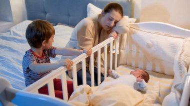 Küçük çocuk yorgun moteher ile gece yatak odasında bebek beşiği sallıyor. Çocuklar ailelere yardım ediyor. Annelik ve uykusuz geceler.