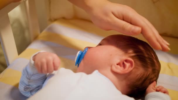 Closeup of young caring mother gently stroking her newborn baby boy lying in crib at night. Concept of happy parenting and family happiness — Stock Video