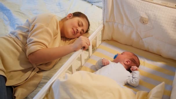 Joven madre cansada se quedó dormida mientras mecía a su hijo en la cuna a altas horas de la noche. Maternidad y noches sin dormir. — Vídeos de Stock