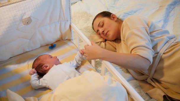 Bezwzględny noworodek leżący w łóżeczku i patrzący na zmęczoną kobietę śpiącą w nocy w łóżku — Wideo stockowe