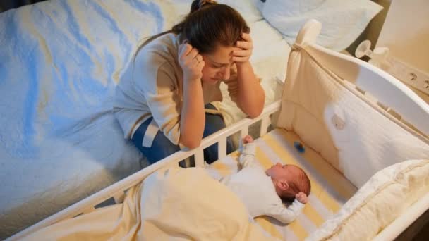 Young tired mother suffering from depression lookin on her sleepless newborn baby in crib at night. Maternal depression after childbirth and sleepless nights. — Stock Video