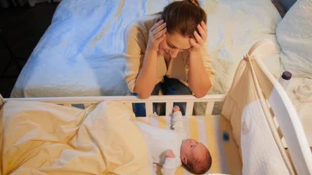 Uykusuz yeni doğmuş bebeğinin beşiğini sallayan yorgun genç bir kadın. Doğum sonrası annelik depresyonu ve uykusuz geceler. — Stok video
