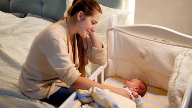 Geceleri gülümseyen güzel bir anne, küçük oğlunun beşiğini sallıyor. Mutlu ebeveynlik ve aile mutluluğu kavramı