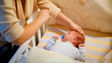 Bebek beşiğinin yanında oturan ve yeni doğmuş bebeğini beşiğinde okşayan şefkatli bir anne. Mutlu ebeveynlik ve aile mutluluğu kavramı