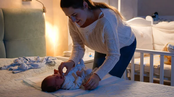 Beautiful smiling mother holding and cuddling her little newborn baby boy at night in bedroom. Mother changes clothes to little child. Concept of loving and caring parents, newborn babies and family — Stock Photo, Image