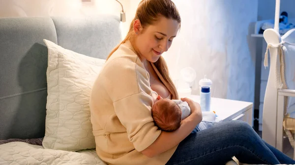 Zâmbind o femeie tânără care se uită și își îmbrățișează bebelușul nou-născut suptând sânii și mâncând lapte noaptea. Conceptul de nutriție sănătoasă și naturală pentru copii. Sănătatea mamei și a copilului. Fericirea familiei — Fotografie, imagine de stoc