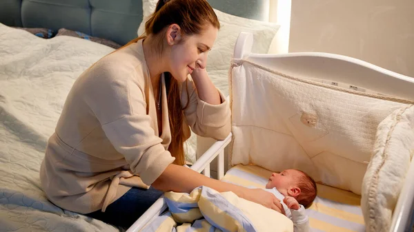 Hermosa joven madre sonriente mecedora cuna de su pequeño hijo por la noche. Concepto de paternidad feliz y felicidad familiar —  Fotos de Stock