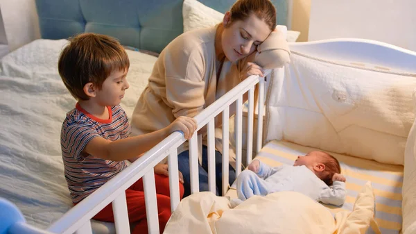 Older boy helping his tired mother rocking baby ccrib in bedroom at night. Children helping parents. Motherhood and sleepless nights — Stock Photo, Image