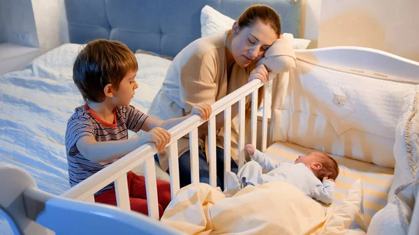 Liten pojke med trött moteher gungande krubba nyfödd pojke i sovrummet på natten. Barn som hjälper föräldrar. Moderskap och sömnlösa nätter. — Stockfoto