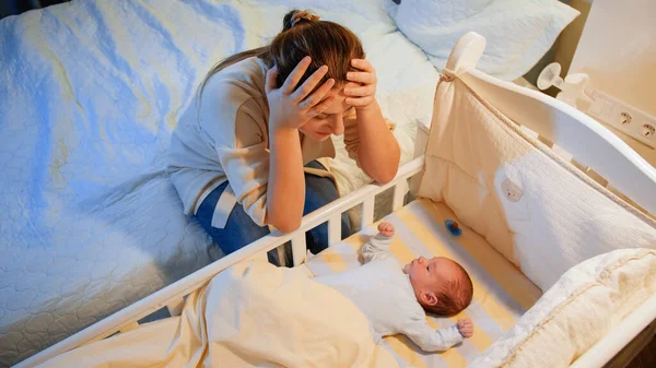 Mladá unavená matka trpí depresí, dívá se na své nespící novorozeně v postýlce v noci. Mateřská deprese po porodu a bezesných nocích. — Stock fotografie