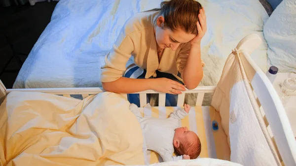 Виснажена молода жінка гойдається на ліжечку свого безсонного новонародженого малюка. материнська депресія після пологів і безсонних ночей . — стокове фото