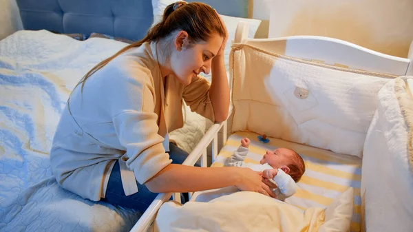 A fiatal, boldog anya ringatja a babáját a bölcsőben, és gyengéden simogatja a kezét. A boldog szülőség és a családi boldogság fogalma — Stock Fotó