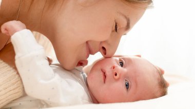 Sevimli yeni doğmuş erkek bebek güneşli bir sabahta büyük bir pencereye karşı genç annesiyle birlikte yatakta yatıyor. Aile mutluluğu ve küçük çocuklu sevgi dolu ebeveynler.