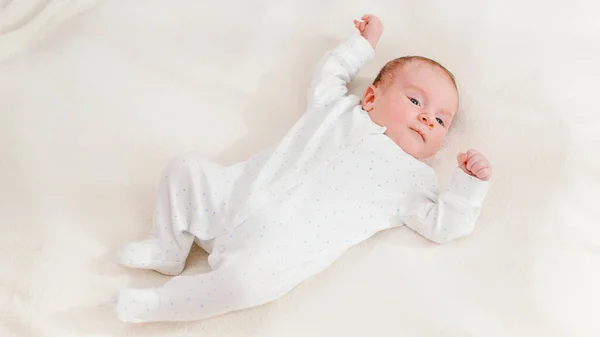 Вид сверху на восхитительного новорожденного в белом костюме, лежащего на кровати и смотрящего на яркое солнце. Новорожденные дети дома — стоковое фото