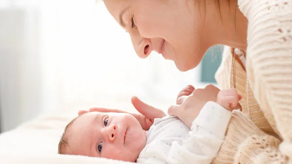 Portrait of adorable smiling newborn baby and happy young mother lying on bed against big window in bedroom. Concept of family happiness and loving parents with little children — Stock Photo, Image