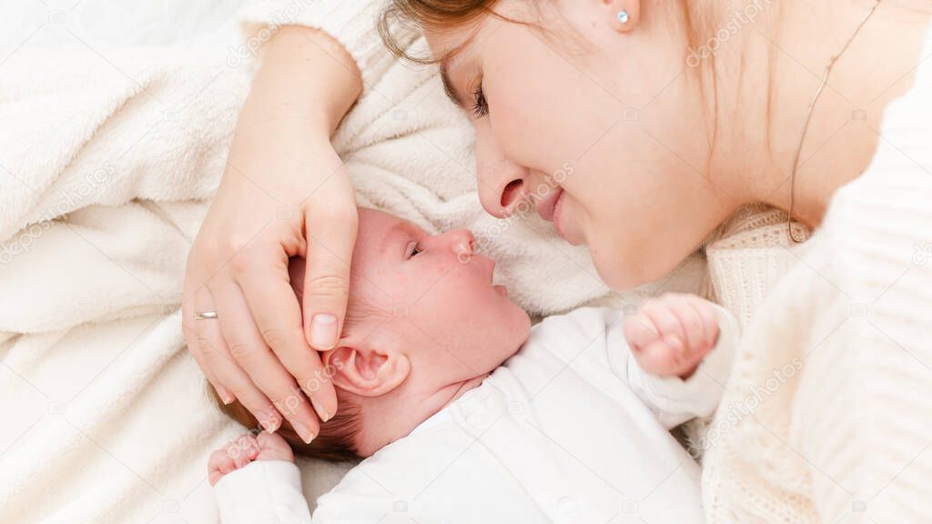 Closeup shot of little newborn baby lying next to mother and crying. Happy smiling woman looking at her little child. Concept of family happiness and loving parents with little children