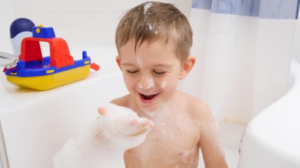 Powolny ruch wesołego śmiejącego się chłopca biorącego kąpiel i dmuchającego piankę mydlaną z ręki w kamerze. Pojęcie higieny dzieci i opieki zdrowotnej w domu. Dzieci dobrze się bawią i bawią — Wideo stockowe