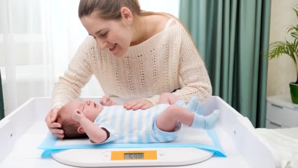 Smiling mother looking and stroking her little son while weighing him on digital scales on changing table at home. Concept of babies and newborn hygiene and healthcare. Caring parents with little — Stock Video