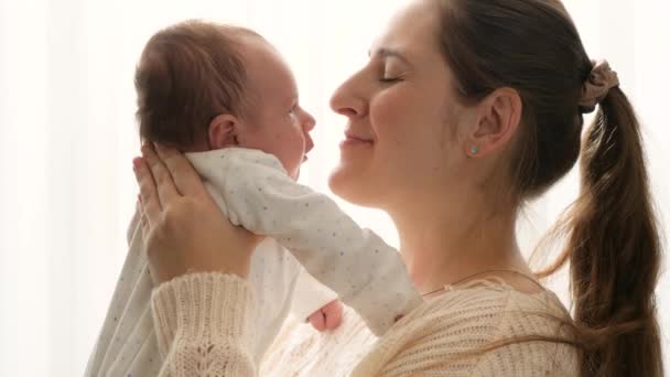 Retrato de la feliz madre sonriente sosteniendo y acariciando a su pequeño bebé recién nacido contra el sol brillante que brilla a través de la ventana en casa. Cuidar a los padres con niños pequeños. — Vídeos de Stock