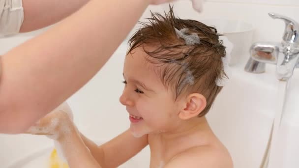 Młoda matka myje swojego synka w wannie i robi mu stylową fryzurę z pianką mydlaną. Pojęcie higieny dzieci i opieki zdrowotnej w domu. Rodzina dobrze się bawi i gra. — Wideo stockowe