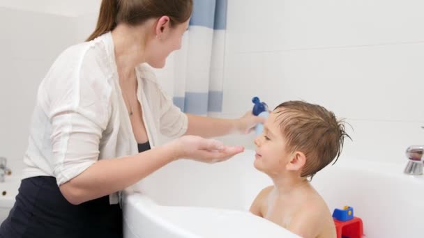 Mladá usměvavá matka si nalila šampon na ruku a umyla vlasy svého malého syna sedícího ve vaně. Koncept dětské hygieny a zdravotní péče doma. Rodina mít čas spolu a hrát si doma — Stock video